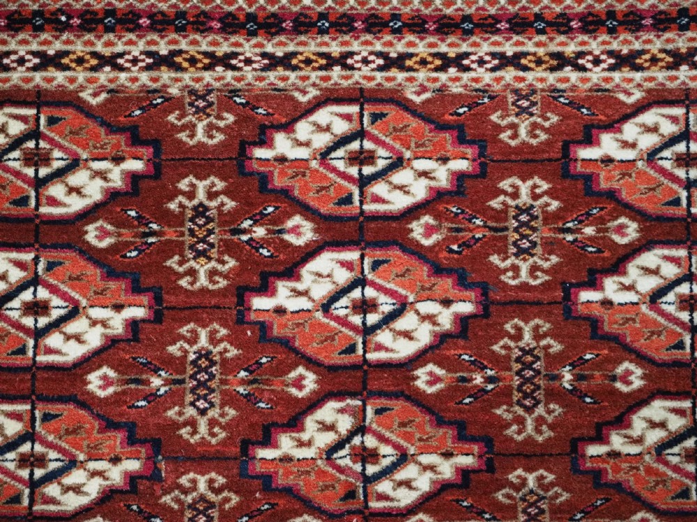 A rectangular handwoven Tekke Turkmen wool rug featuring repeating primary gul motifs in deep red, navy, terracotta, and ivory tones surrounded by geometric borders. View VII