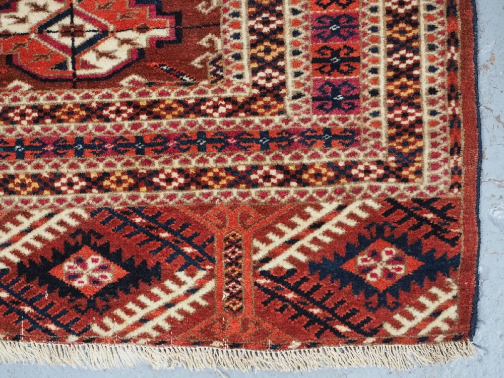A rectangular handwoven Tekke Turkmen wool rug featuring repeating primary gul motifs in deep red, navy, terracotta, and ivory tones surrounded by geometric borders. View X