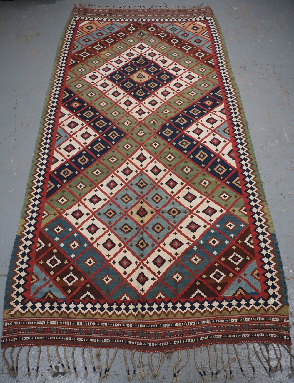 An antique Persian Qashqai tribal kilim rug featuring a dense geometric diamond lattice pattern in terracotta, navy blue, sage green, and cream wool with fringed ends.