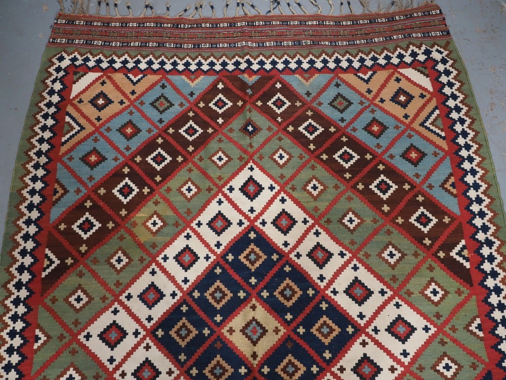 An antique Persian Qashqai tribal kilim rug featuring a dense geometric diamond lattice pattern in terracotta, navy blue, sage green, and cream wool with fringed ends. View III