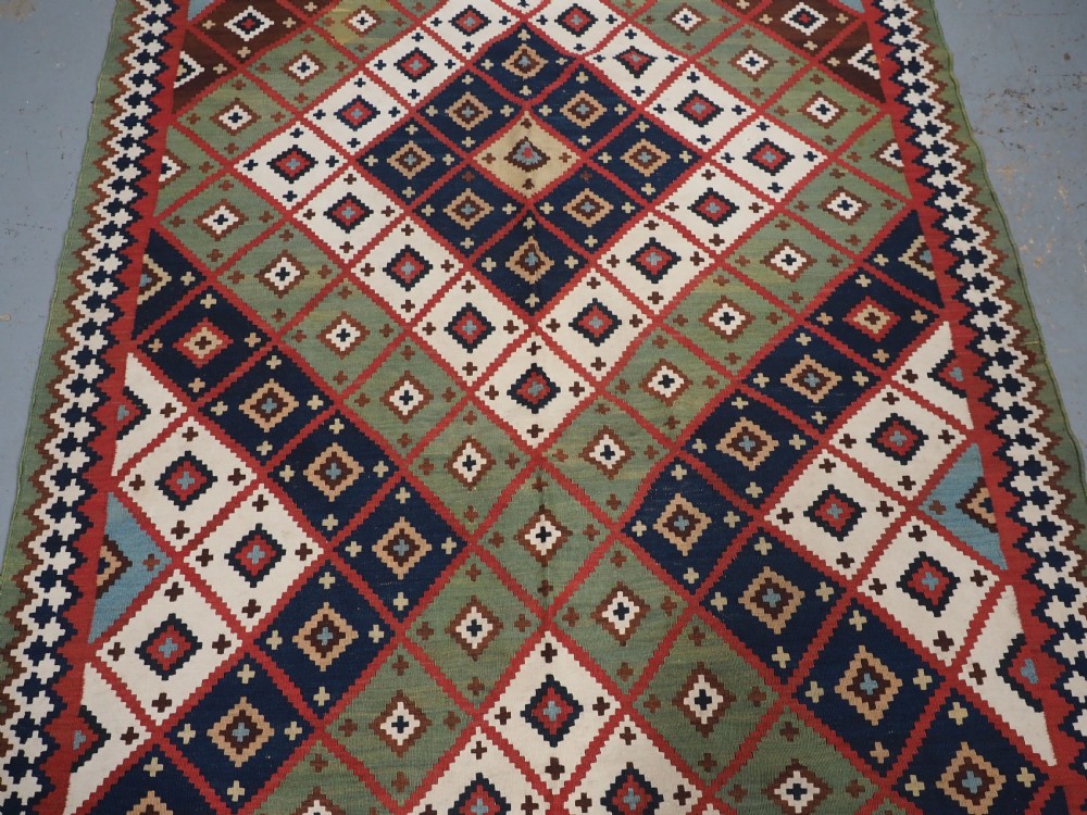An antique Persian Qashqai tribal kilim rug featuring a dense geometric diamond lattice pattern in terracotta, navy blue, sage green, and cream wool with fringed ends. View IV