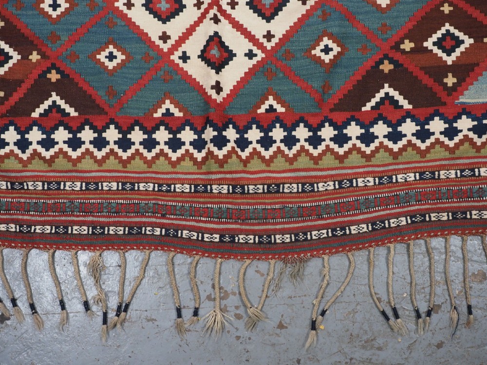 An antique Persian Qashqai tribal kilim rug featuring a dense geometric diamond lattice pattern in terracotta, navy blue, sage green, and cream wool with fringed ends. View X