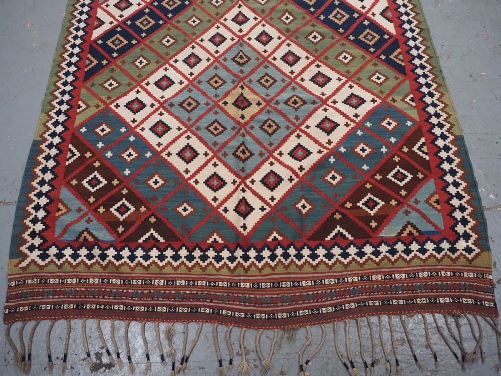 An antique Persian Qashqai tribal kilim rug featuring a dense geometric diamond lattice pattern in terracotta, navy blue, sage green, and cream wool with fringed ends. View XI