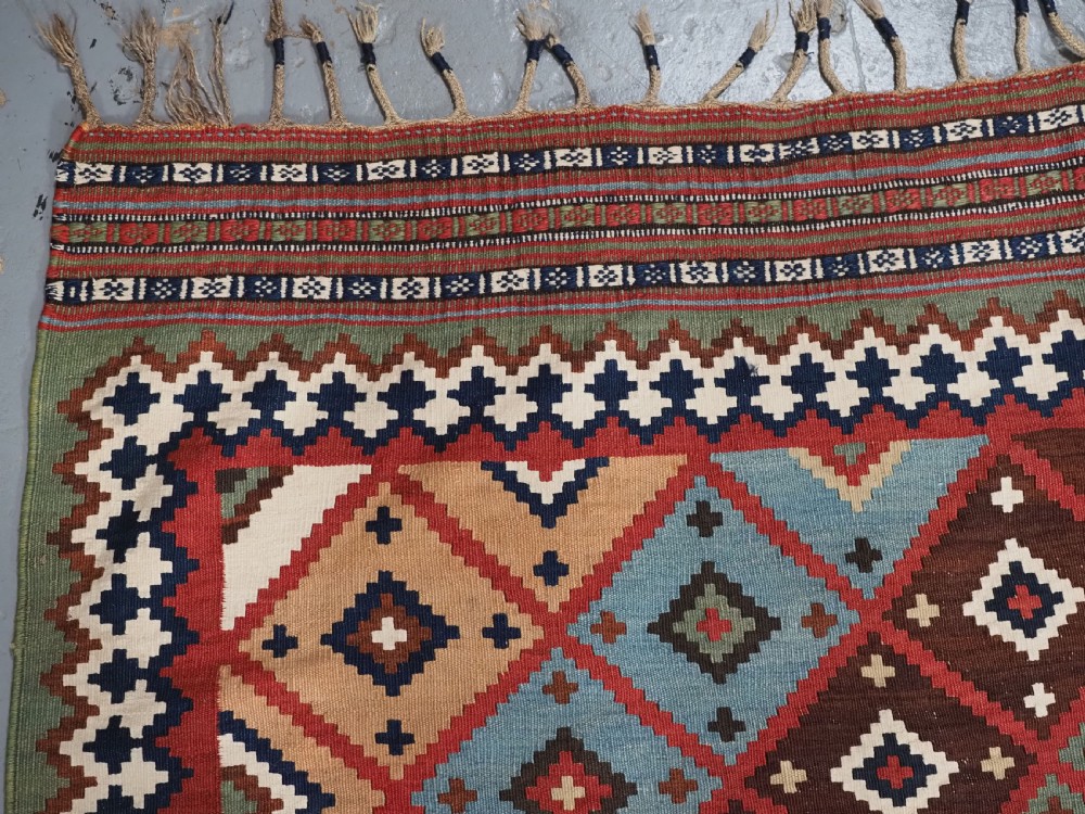 An antique Persian Qashqai tribal kilim rug featuring a dense geometric diamond lattice pattern in terracotta, navy blue, sage green, and cream wool with fringed ends. View XIII