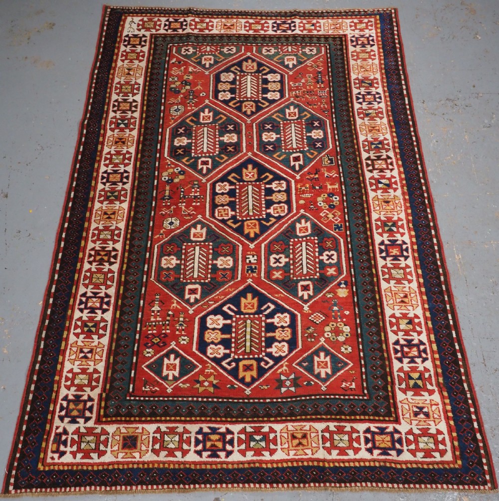 An antique hand-knotted Caucasian rug featuring geometric diamond medallions and folk art figures on a deep red wool field.