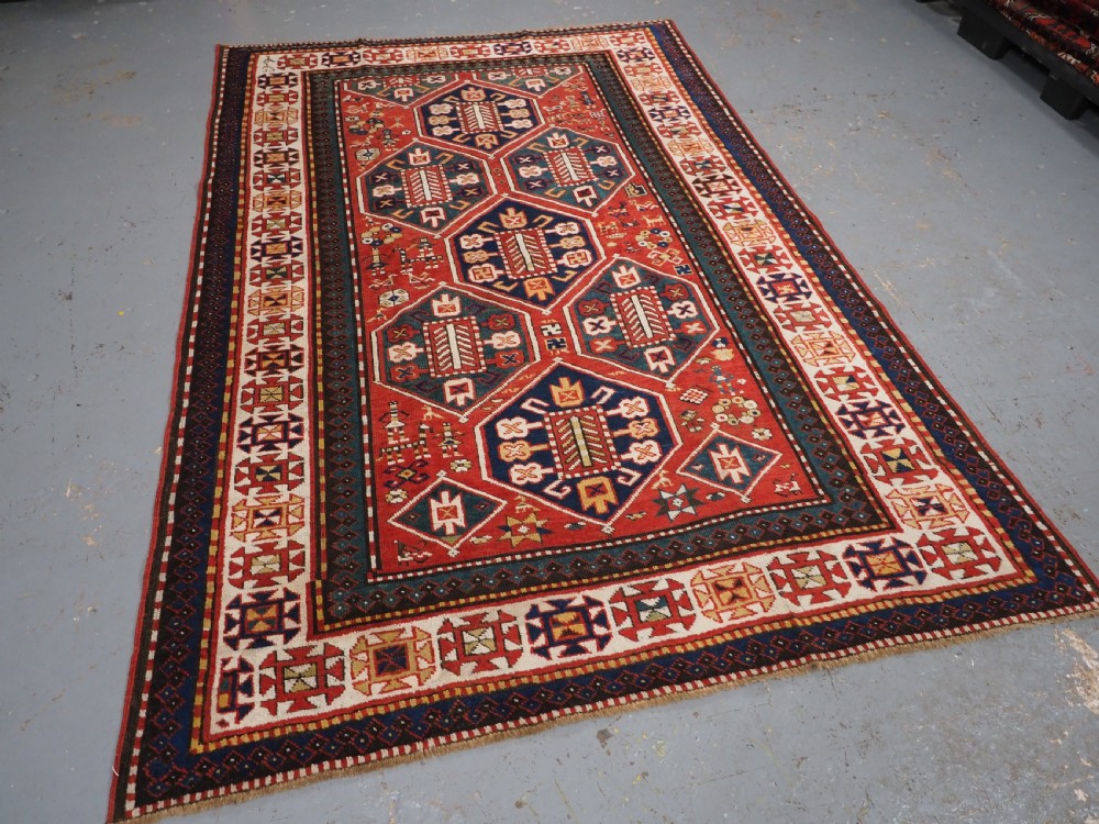 An antique hand-knotted Caucasian rug featuring geometric diamond medallions and folk art figures on a deep red wool field. View II