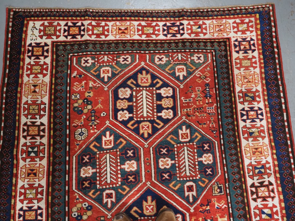 An antique hand-knotted Caucasian rug featuring geometric diamond medallions and folk art figures on a deep red wool field. View III