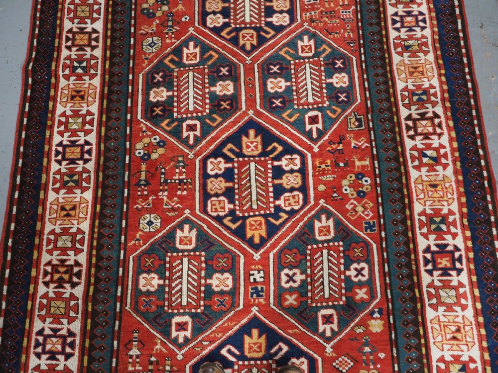 An antique hand-knotted Caucasian rug featuring geometric diamond medallions and folk art figures on a deep red wool field. View IV