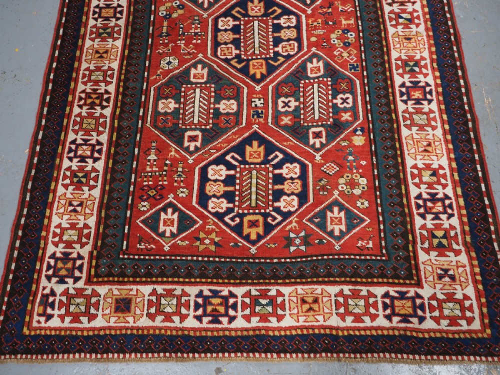 An antique hand-knotted Caucasian rug featuring geometric diamond medallions and folk art figures on a deep red wool field. View V
