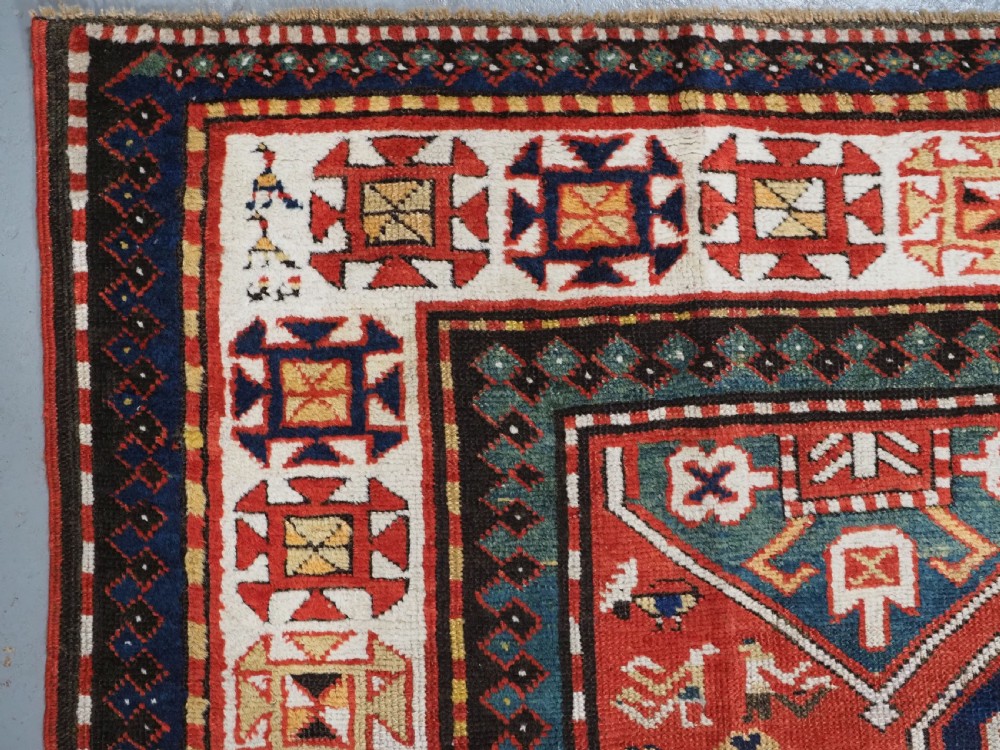 An antique hand-knotted Caucasian rug featuring geometric diamond medallions and folk art figures on a deep red wool field. View VI
