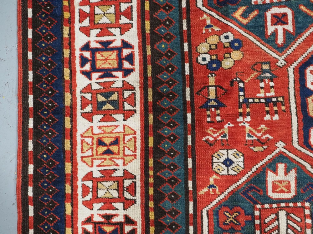 An antique hand-knotted Caucasian rug featuring geometric diamond medallions and folk art figures on a deep red wool field. View VII