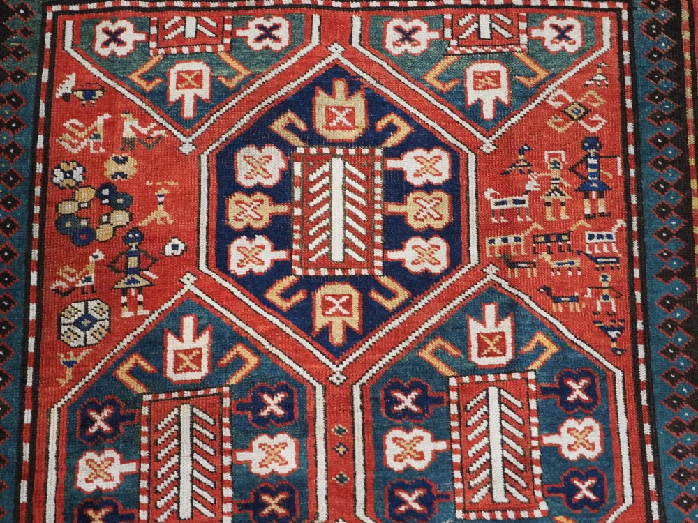 An antique hand-knotted Caucasian rug featuring geometric diamond medallions and folk art figures on a deep red wool field. View VIII
