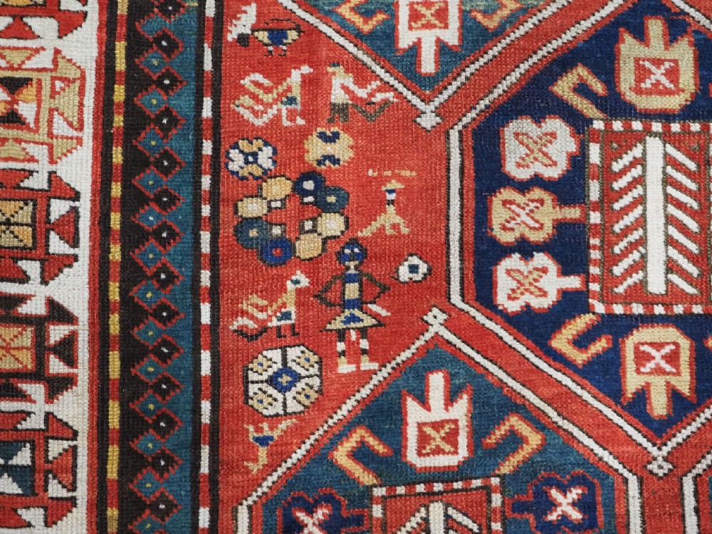 An antique hand-knotted Caucasian rug featuring geometric diamond medallions and folk art figures on a deep red wool field. View IX