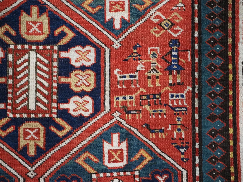An antique hand-knotted Caucasian rug featuring geometric diamond medallions and folk art figures on a deep red wool field. View X