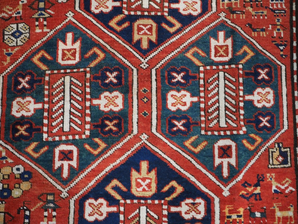 An antique hand-knotted Caucasian rug featuring geometric diamond medallions and folk art figures on a deep red wool field. View XI