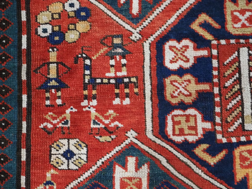 An antique hand-knotted Caucasian rug featuring geometric diamond medallions and folk art figures on a deep red wool field. View XII