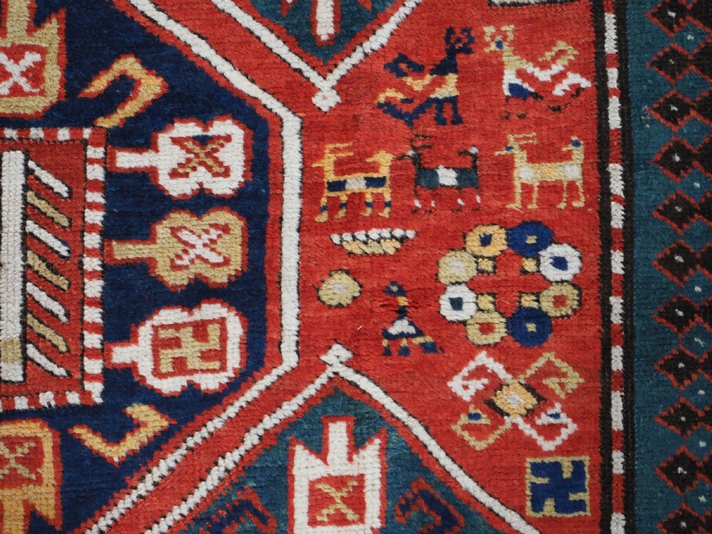 An antique hand-knotted Caucasian rug featuring geometric diamond medallions and folk art figures on a deep red wool field. View XIII