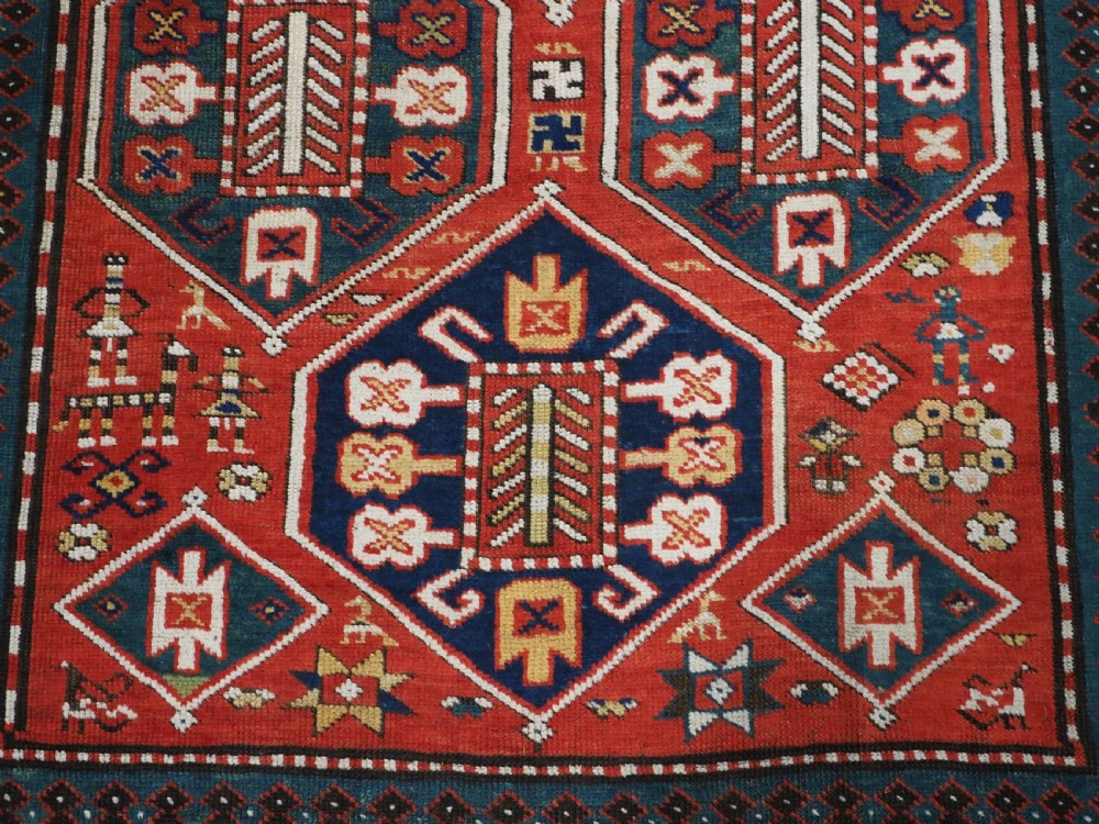 An antique hand-knotted Caucasian rug featuring geometric diamond medallions and folk art figures on a deep red wool field. View XIV