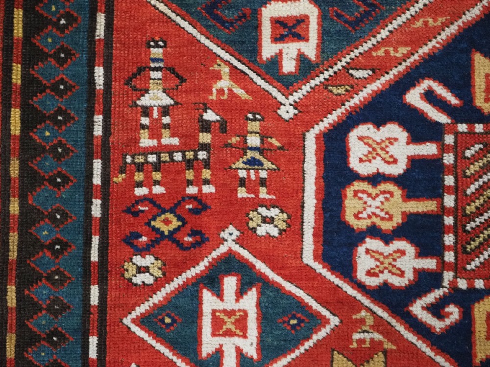 An antique hand-knotted Caucasian rug featuring geometric diamond medallions and folk art figures on a deep red wool field. View XV