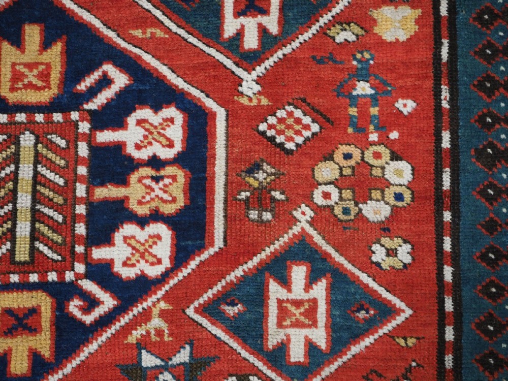 An antique hand-knotted Caucasian rug featuring geometric diamond medallions and folk art figures on a deep red wool field. View XVI