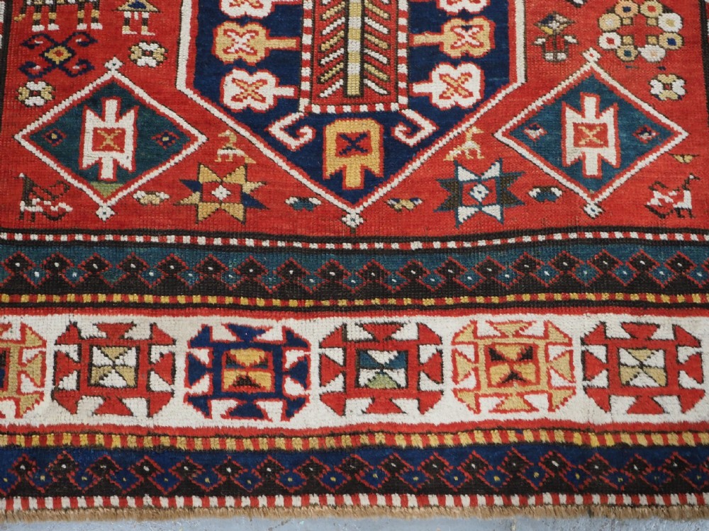 An antique hand-knotted Caucasian rug featuring geometric diamond medallions and folk art figures on a deep red wool field. View XVII
