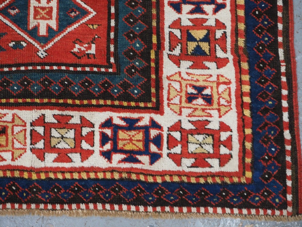 An antique hand-knotted Caucasian rug featuring geometric diamond medallions and folk art figures on a deep red wool field. View XVIII