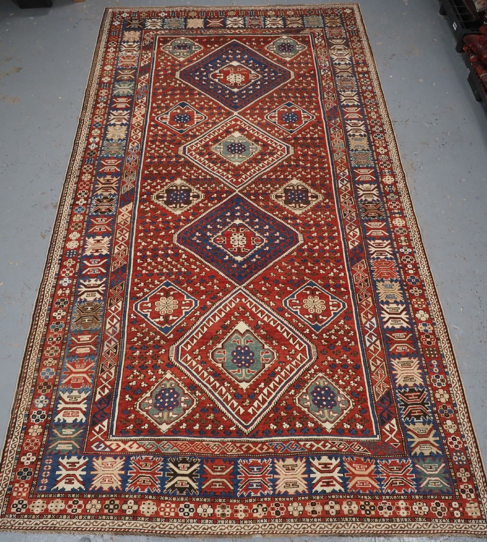 An antique hand-knotted Caucasian Shirvan Baku Khila wool rug featuring geometric diamond medallions on a red ground with navy blue and ivory borders.