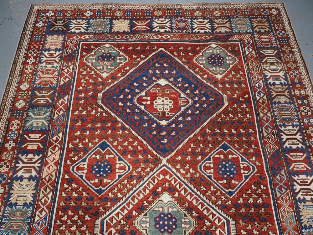 An antique hand-knotted Caucasian Shirvan Baku Khila wool rug featuring geometric diamond medallions on a red ground with navy blue and ivory borders. View III
