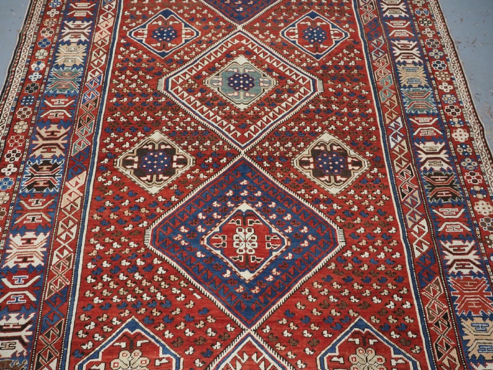 An antique hand-knotted Caucasian Shirvan Baku Khila wool rug featuring geometric diamond medallions on a red ground with navy blue and ivory borders. View IV