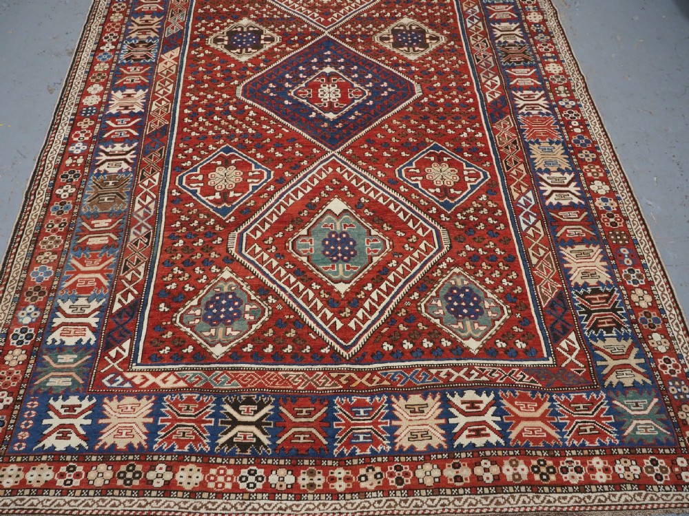 An antique hand-knotted Caucasian Shirvan Baku Khila wool rug featuring geometric diamond medallions on a red ground with navy blue and ivory borders. View V