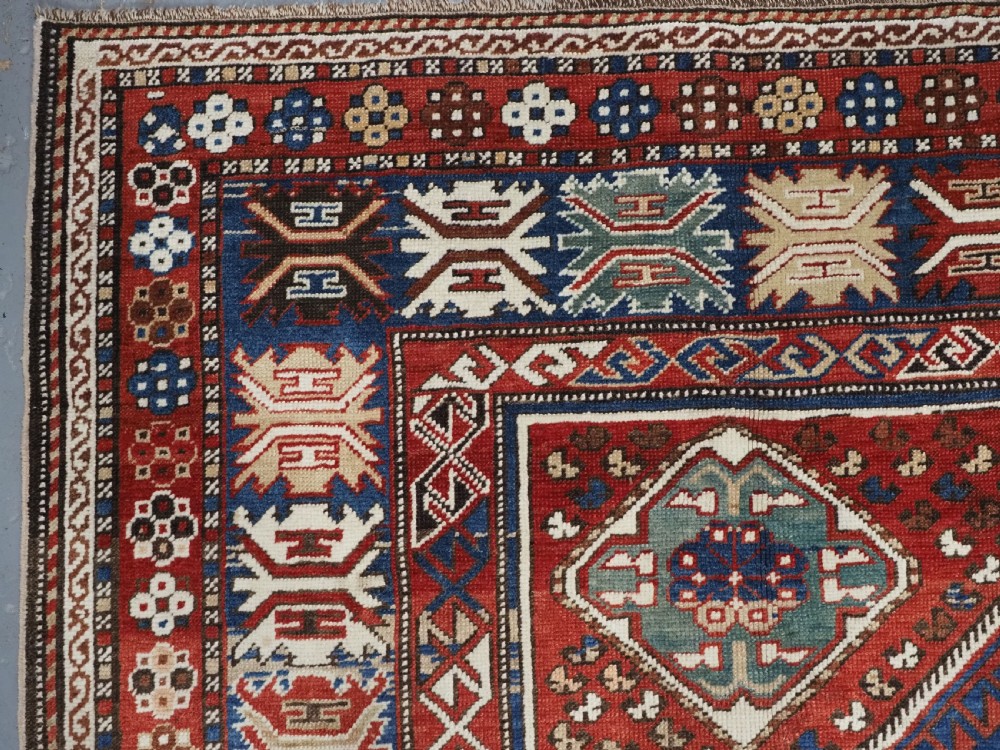An antique hand-knotted Caucasian Shirvan Baku Khila wool rug featuring geometric diamond medallions on a red ground with navy blue and ivory borders. View VI