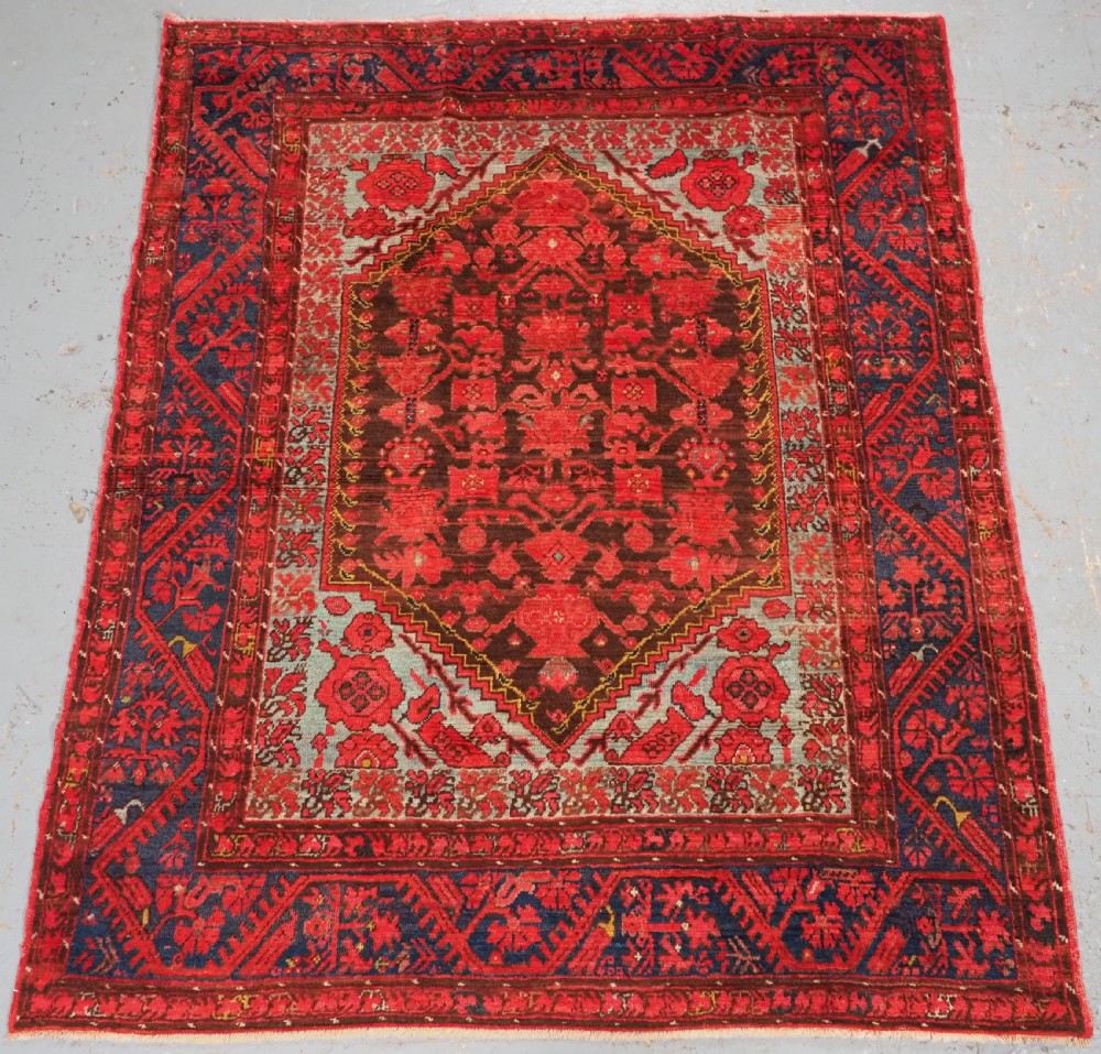 An antique hand-knotted Turkish Komurcu Kula wool rug featuring a central diamond medallion in crimson red, deep blue, dark brown, and grey-blue tones.