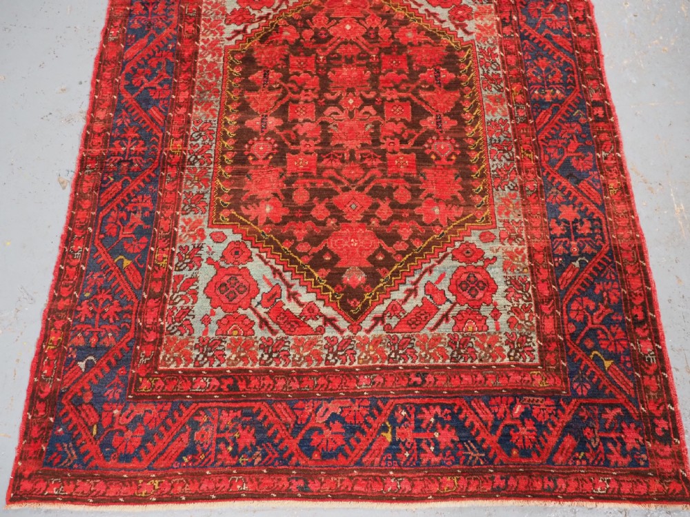 An antique hand-knotted Turkish Komurcu Kula wool rug featuring a central diamond medallion in crimson red, deep blue, dark brown, and grey-blue tones. View IV