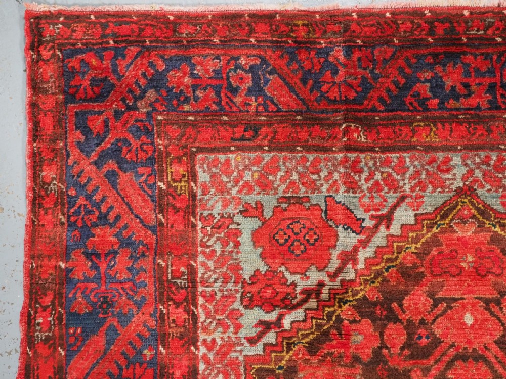 An antique hand-knotted Turkish Komurcu Kula wool rug featuring a central diamond medallion in crimson red, deep blue, dark brown, and grey-blue tones. View V