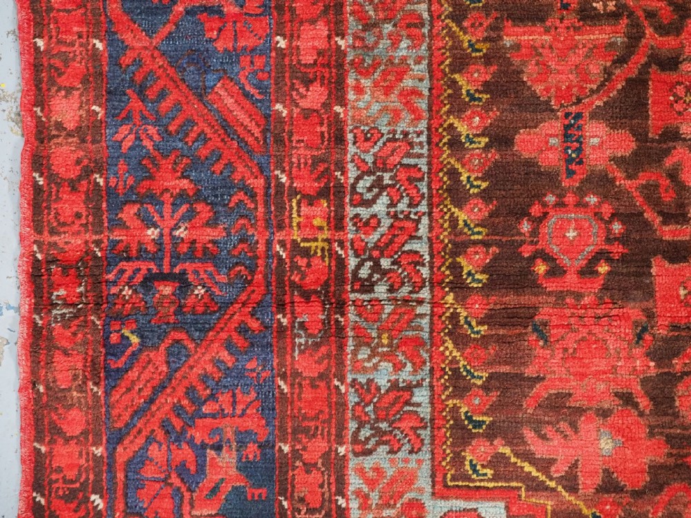 An antique hand-knotted Turkish Komurcu Kula wool rug featuring a central diamond medallion in crimson red, deep blue, dark brown, and grey-blue tones. View VI