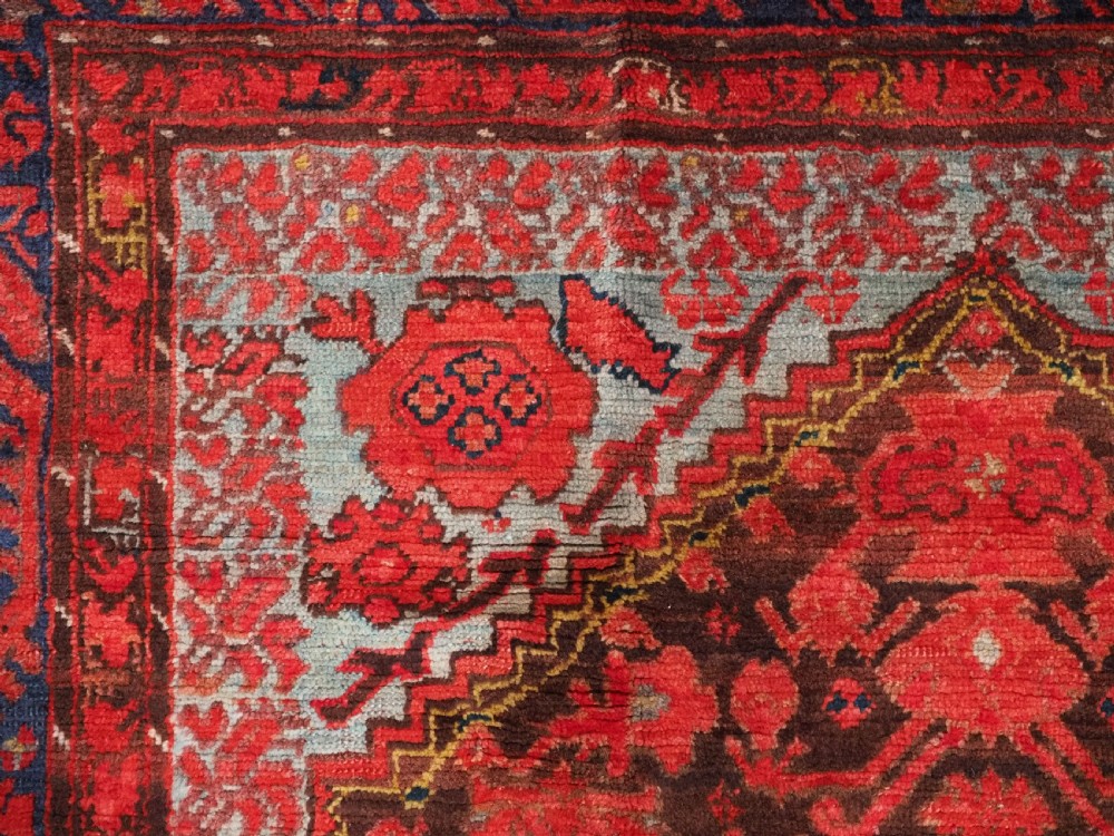 An antique hand-knotted Turkish Komurcu Kula wool rug featuring a central diamond medallion in crimson red, deep blue, dark brown, and grey-blue tones. View VII