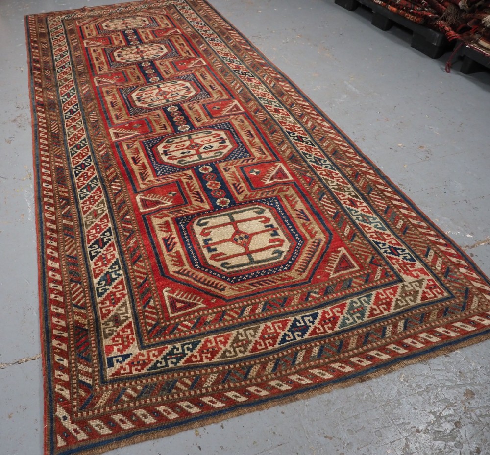 An antique Caucasian Shirvan long wool runner rug featuring terracotta red, navy blue, and beige octagonal geometric medallions within patterned borders. View II