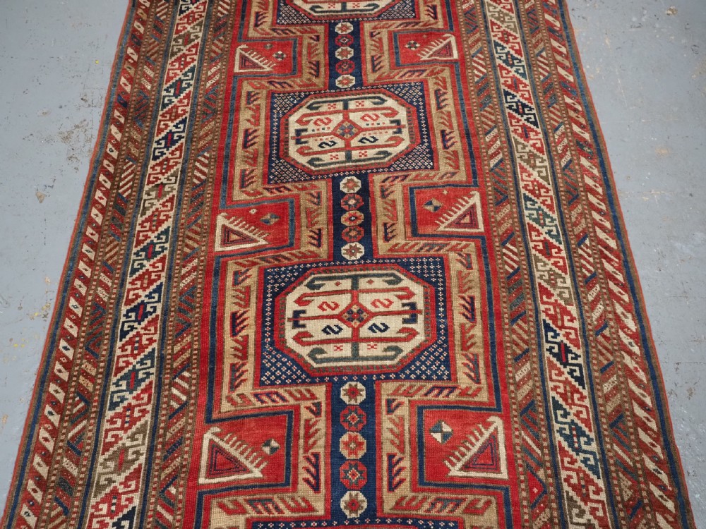 An antique Caucasian Shirvan long wool runner rug featuring terracotta red, navy blue, and beige octagonal geometric medallions within patterned borders. View IV