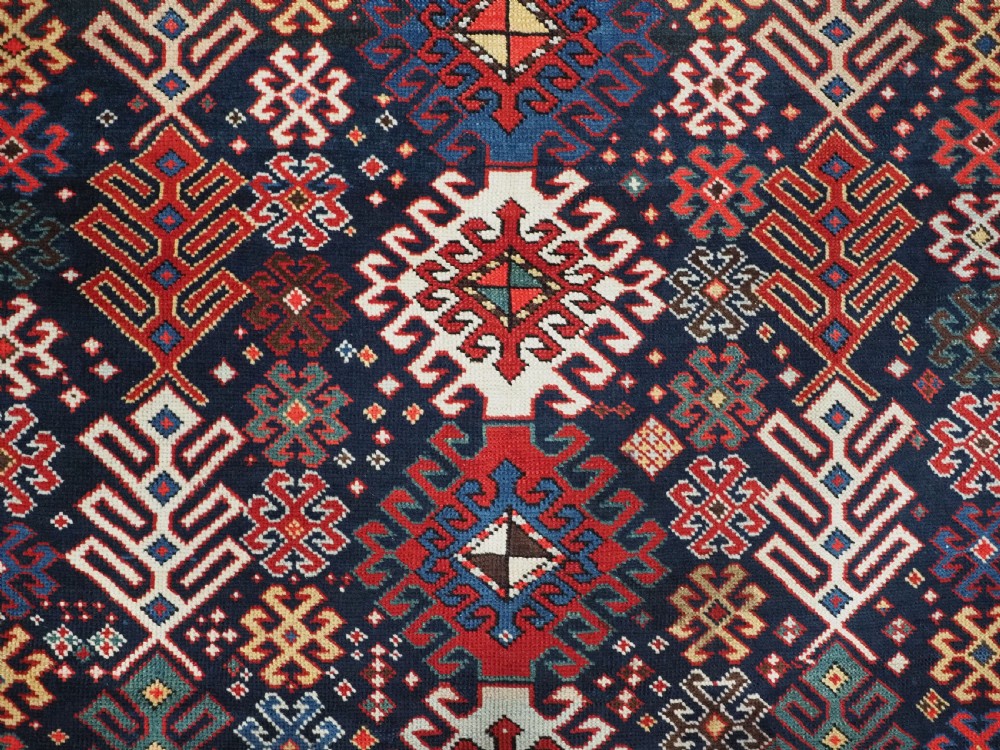 An antique long Caucasian runner rug featuring a dark navy field filled with multi-coloured hooked diamond medallions and detailed geometric borders. View IX