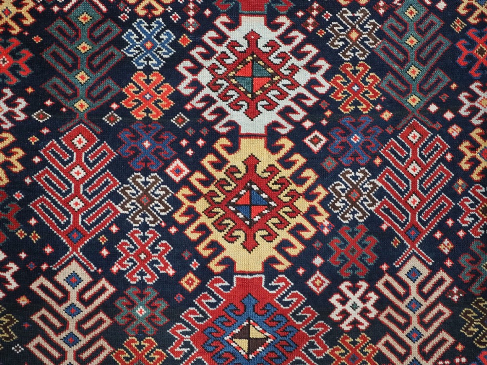 An antique long Caucasian runner rug featuring a dark navy field filled with multi-coloured hooked diamond medallions and detailed geometric borders. View X