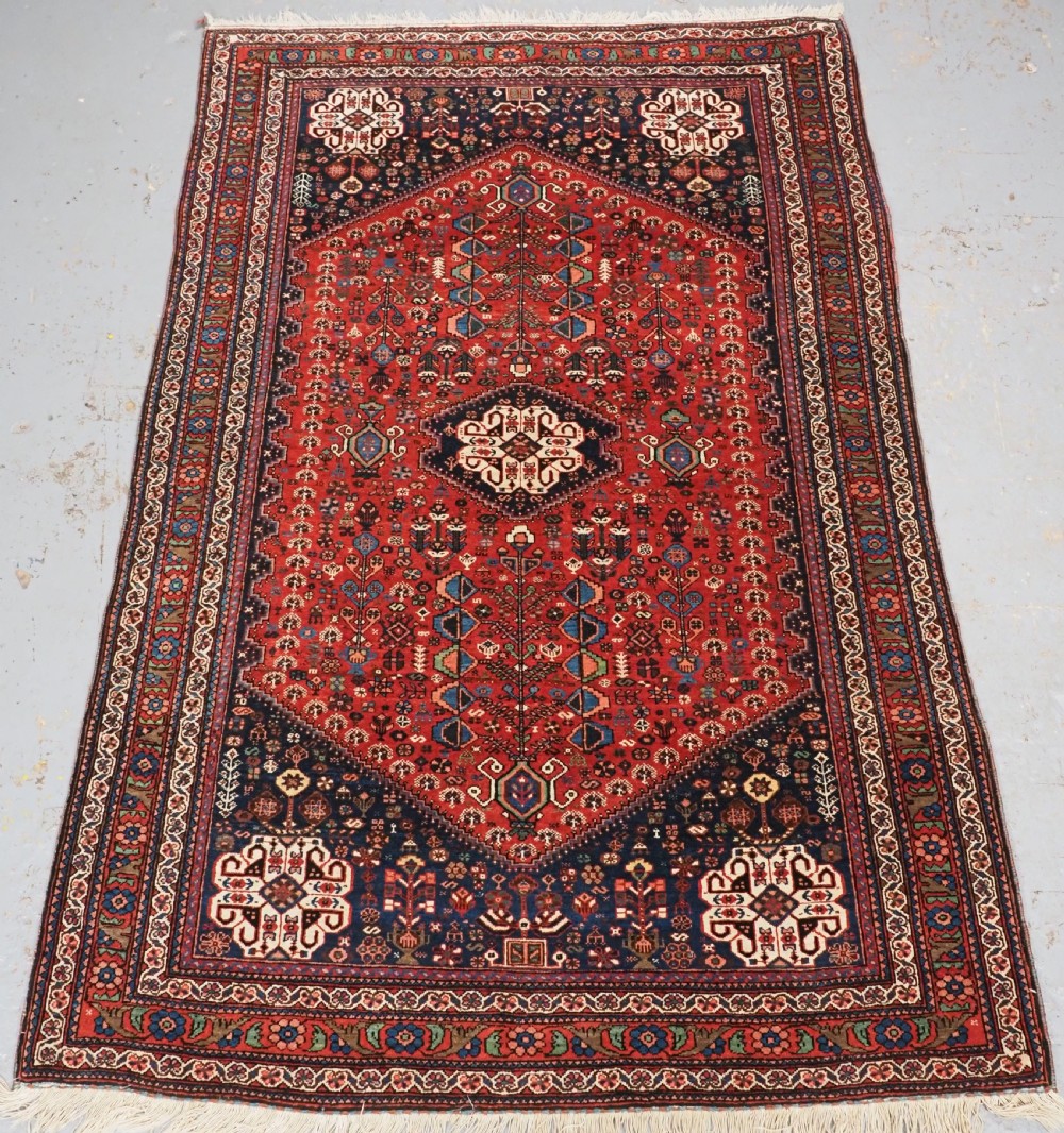 An antique hand-knotted Abadeh wool rug featuring a central diamond medallion, terracotta red field, and navy blue borders with intricate tribal geometric patterns.