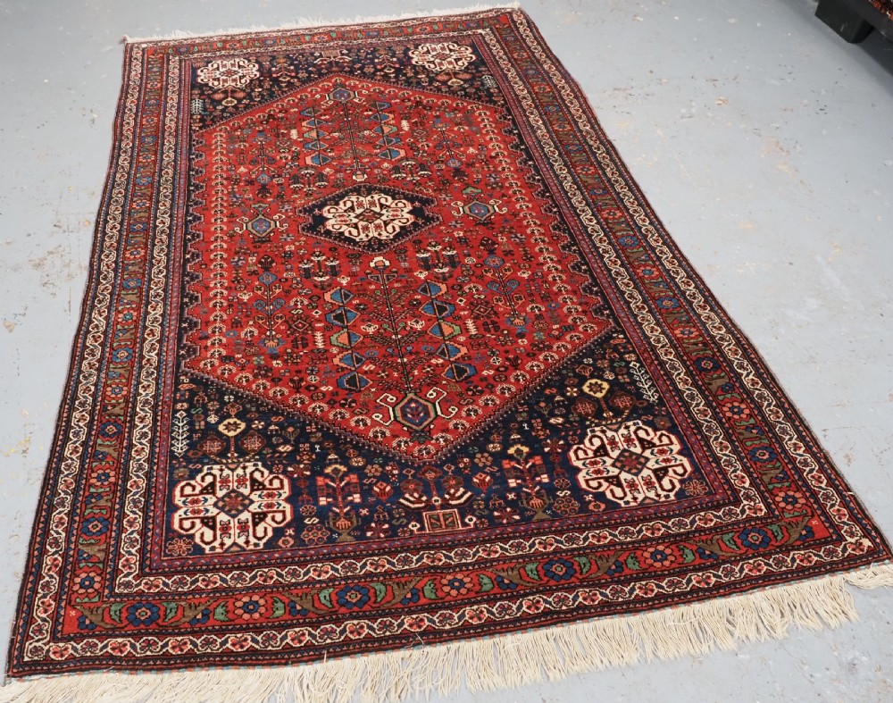 An antique hand-knotted Abadeh wool rug featuring a central diamond medallion, terracotta red field, and navy blue borders with intricate tribal geometric patterns. View II