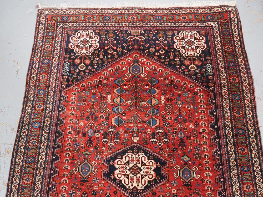 An antique hand-knotted Abadeh wool rug featuring a central diamond medallion, terracotta red field, and navy blue borders with intricate tribal geometric patterns. View III