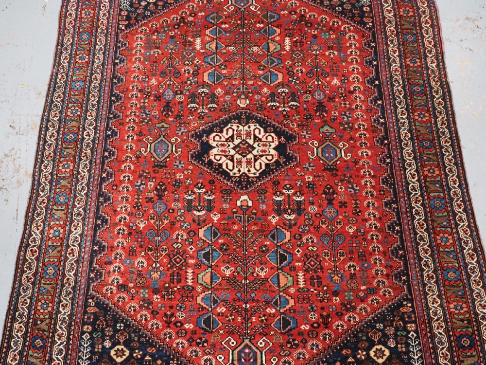 An antique hand-knotted Abadeh wool rug featuring a central diamond medallion, terracotta red field, and navy blue borders with intricate tribal geometric patterns. View IV