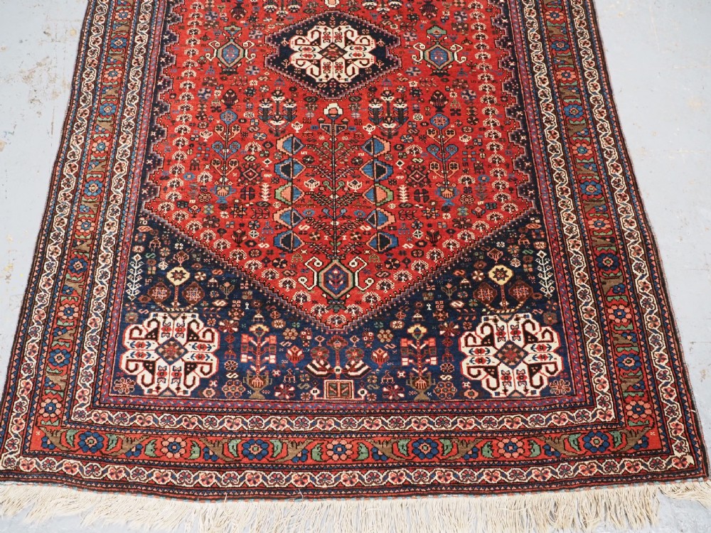 An antique hand-knotted Abadeh wool rug featuring a central diamond medallion, terracotta red field, and navy blue borders with intricate tribal geometric patterns. View V
