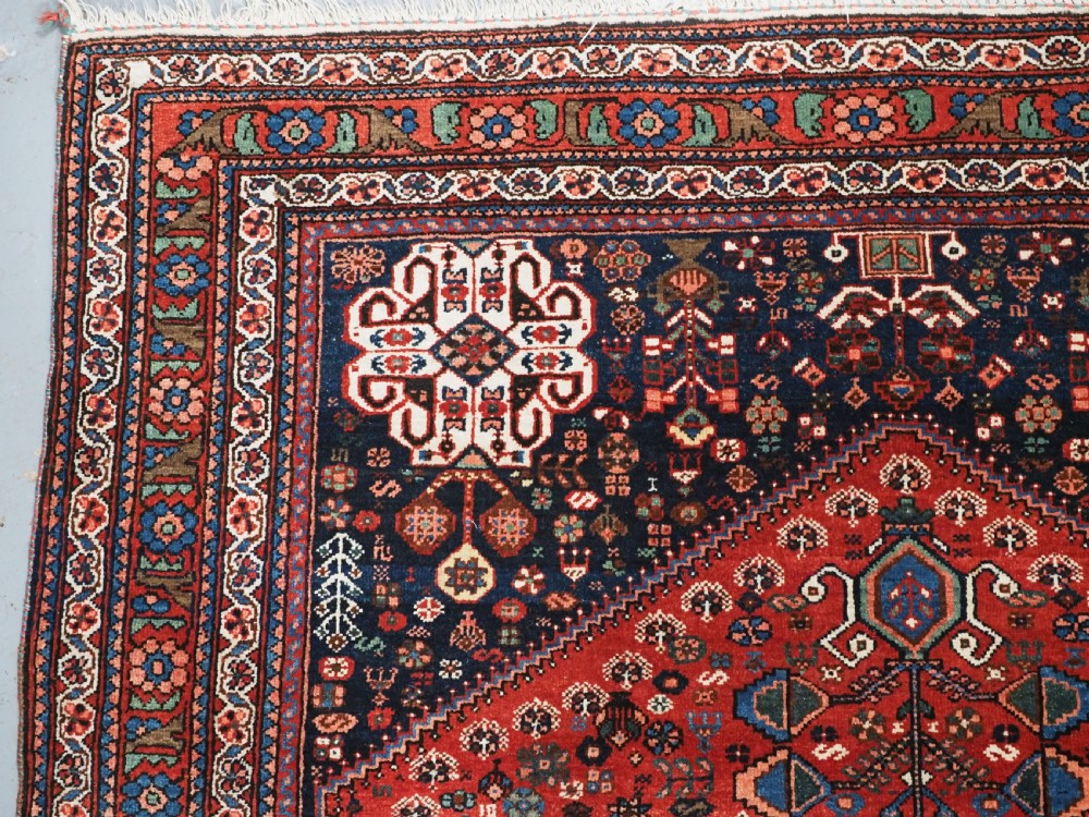 An antique hand-knotted Abadeh wool rug featuring a central diamond medallion, terracotta red field, and navy blue borders with intricate tribal geometric patterns. View VI