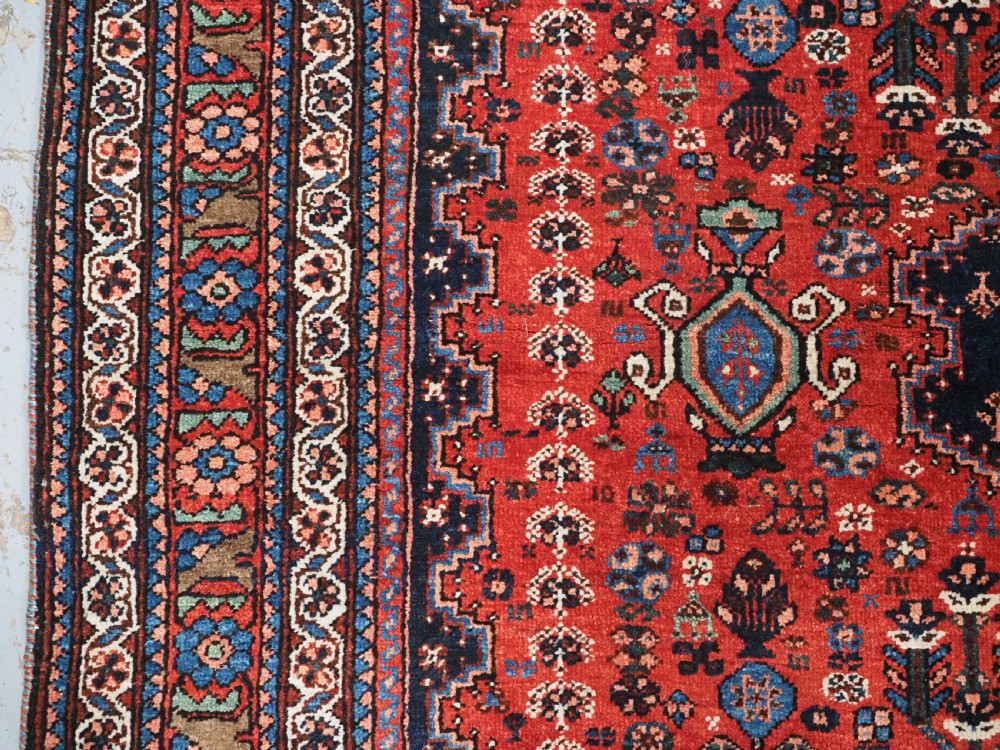 An antique hand-knotted Abadeh wool rug featuring a central diamond medallion, terracotta red field, and navy blue borders with intricate tribal geometric patterns. View VII