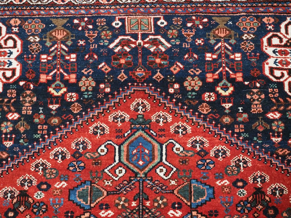 An antique hand-knotted Abadeh wool rug featuring a central diamond medallion, terracotta red field, and navy blue borders with intricate tribal geometric patterns. View IX