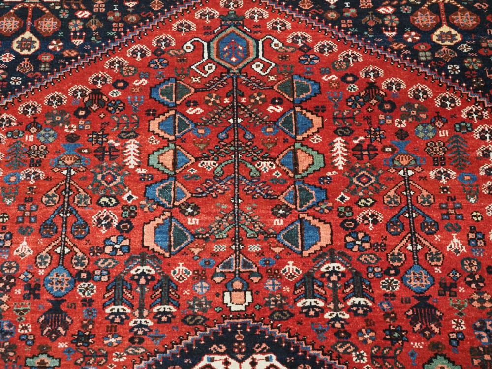 An antique hand-knotted Abadeh wool rug featuring a central diamond medallion, terracotta red field, and navy blue borders with intricate tribal geometric patterns. View X