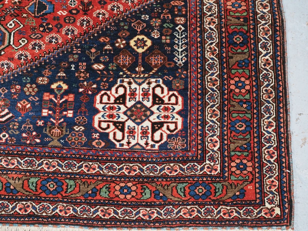 An antique hand-knotted Abadeh wool rug featuring a central diamond medallion, terracotta red field, and navy blue borders with intricate tribal geometric patterns. View XIII